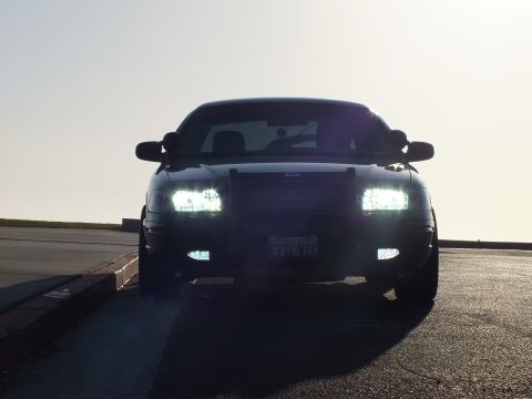 2003 Ford Crown Victoria - Front view of a black four-door with brush guard, headlights on, sun flare crossing the roof.