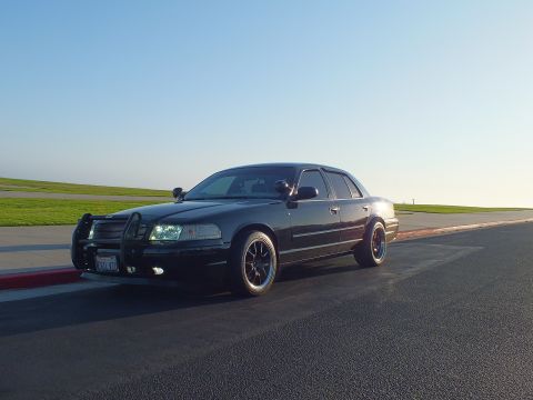 2003 Ford Crown Victoria - Front-quarter view of a blacked-out sedan with push bar parked on pavement, ocean horizon behind.