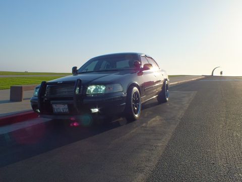2003 Ford Crown Victoria - Backlit front-quarter view of a black four-door with push bar, sculpture arch in the distance.