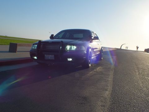 2003 Ford Crown Victoria - Front-quarter of a black sedan with brush guard and lights lit, sun flare across the pavement.
