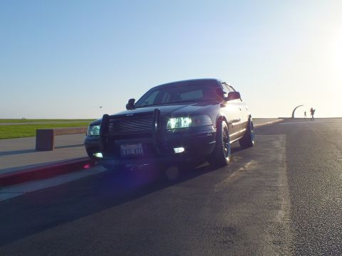 2003 Ford Crown Victoria - Front-quarter view of a black four-door with grille guard, sun behind it and a sculpture arch right.