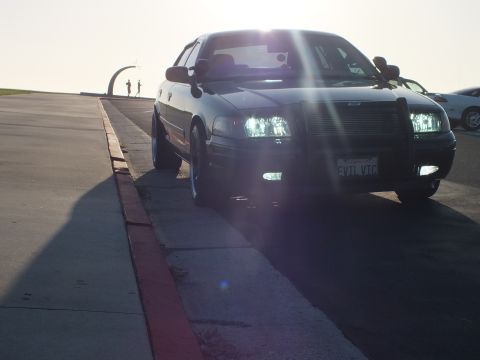 2003 Ford Crown Victoria - Backlit front-quarter of a black sedan parked along a red curb, sun flare and a white car to the right.
