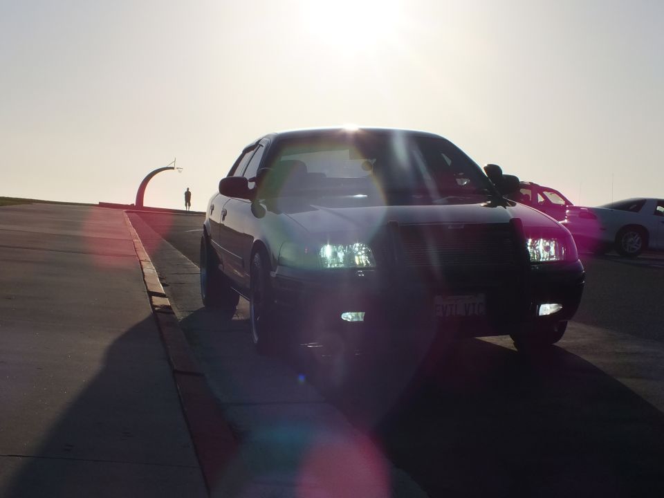 Black sedan front three-quarter with grille guard backlit by sun at coastal overlook