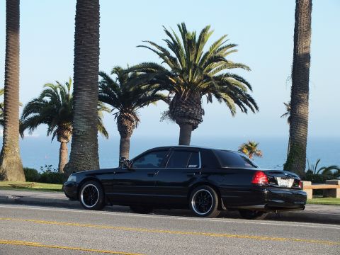 2003 Ford Crown Victoria - Side profile of a blacked-out sedan on a coastal road, palm trees and sea behind it.