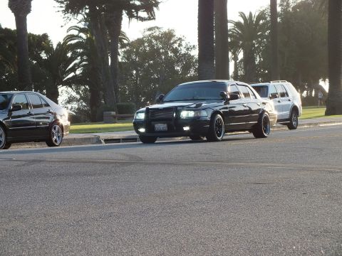 2003 Ford Crown Victoria - Black sedan with push bar angled across a street, other vehicles parked around it.