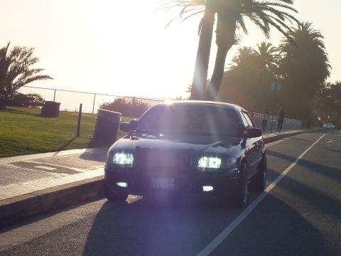 2003 Ford Crown Victoria - Black sedan facing camera with sun flare behind, headlights and fog lamps lit.