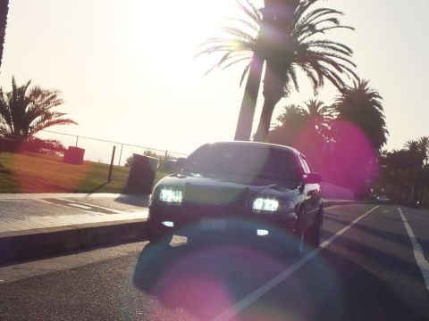 2003 Ford Crown Victoria - Black sedan on roadside washed in sunset glare, headlights illuminated.