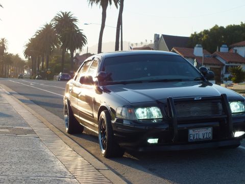2003 Ford Crown Victoria - Crown Victoria at the curb, push bar and headlights lit, palms and tile-roof homes.