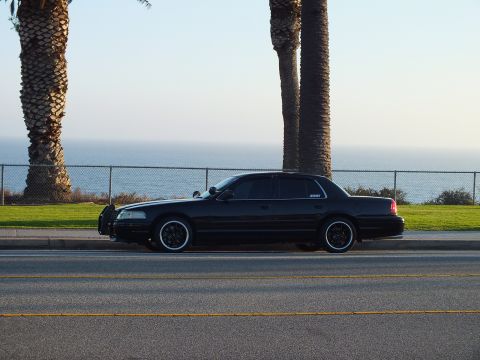 2003 Ford Crown Victoria - Side view of a black sedan on black wheels parked along an oceanfront fence.