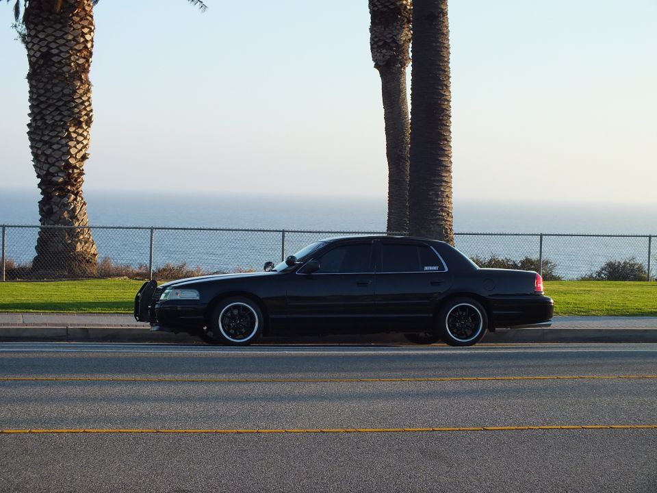 Black full-size sedan side profile by chain-link fence with ocean behind