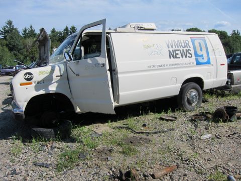 1991 Ford LTD Crown Victoria - A white van, likely a news vehicle, sits in a junkyard with its doors open and hood ajar.