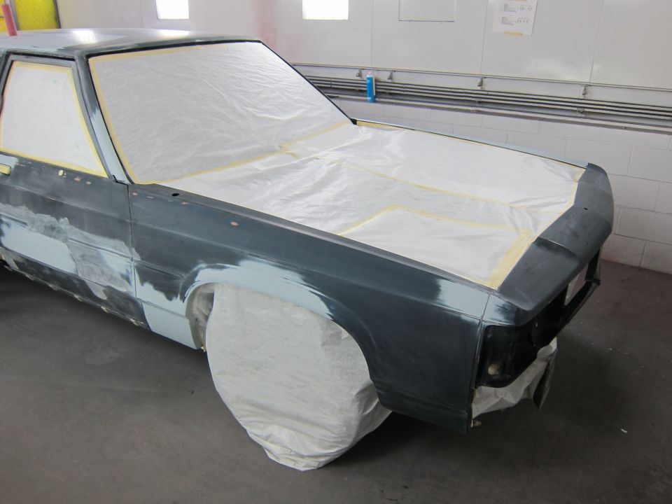Car body covered in plastic sheeting and masking tape, with primer and body filler visible on the fenders and hood. It's in a workshop setting
