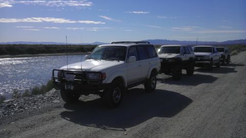 1985 Toyota 4Runner - A line of off-road vehicles, including a Toyota Land Cruiser 80 Series, parked on a dirt road next to a body of water under a clear sky.