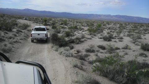 1985 Toyota 4Runner - A white pickup truck travels down a dusty desert road, with another vehicle visible in the distance under a vast sky.
