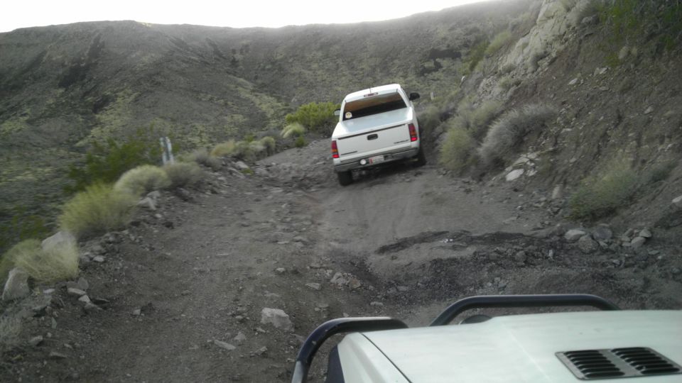 White pickup truck driving down a steep, rocky dirt road in a desert landscape. Another vehicle's interior is visible in the foreground