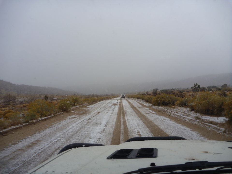 View from inside a vehicle driving on a dirt road covered in snow, with another vehicle ahead