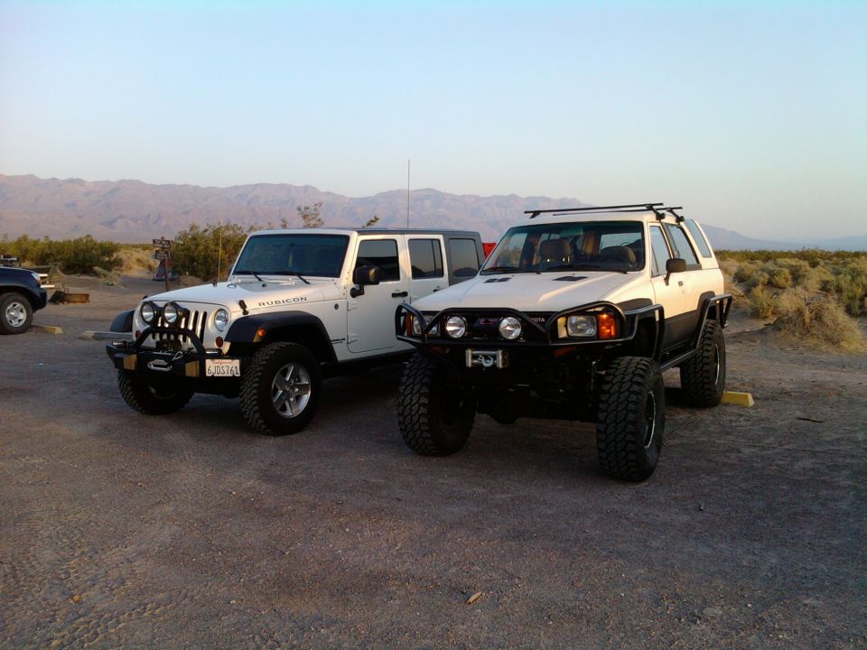 White Jeep Wrangler and a white Toyota 4Runner parked on dirt lot with mountains in background