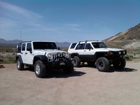 1985 Toyota 4Runner - Two modified SUVs, a Jeep and a Toyota, are parked side-by-side in a dry, mountainous area under a clear blue sky.