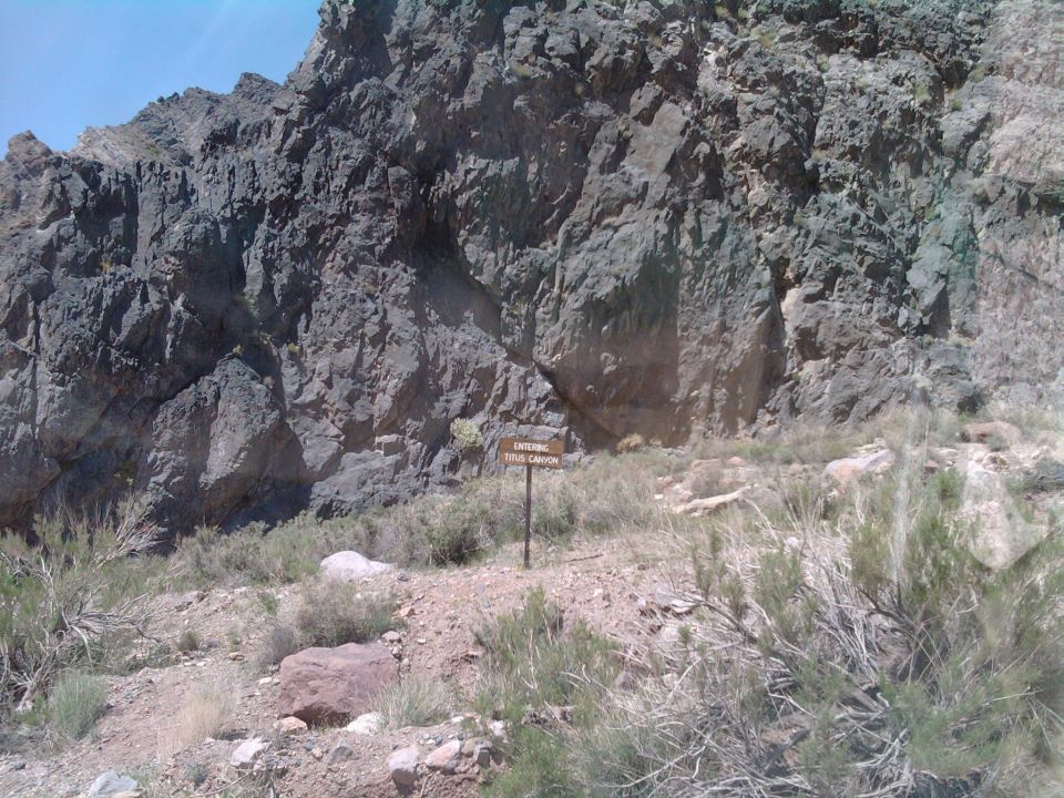Wooden sign reading "Entering Titus Canyon" stands in a dry, rocky landscape with sparse vegetation