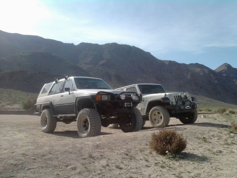 1985 Toyota 4Runner - Two modified off-road vehicles, a Toyota 4Runner and a Jeep Wrangler, are parked side-by-side in a dry, mountainous desert environment.