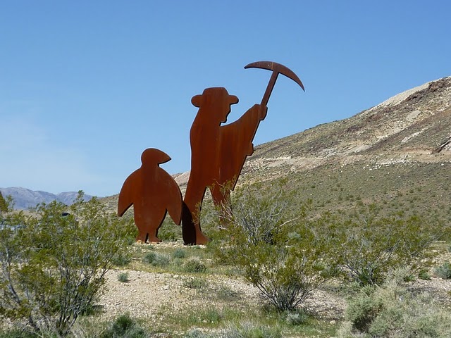 Metal sculpture of a prospector with a pickaxe and a penguin in a desert landscape
