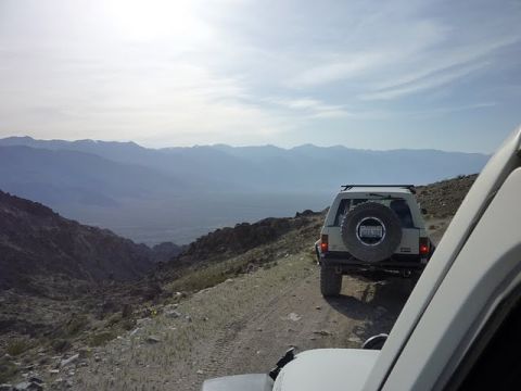 1985 Toyota 4Runner - Two SUVs navigate a dusty mountain trail with vast, layered mountains stretching into the distance under a hazy sky.