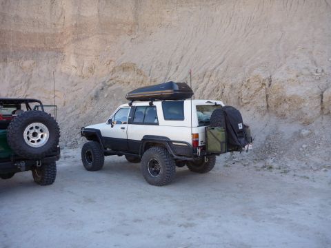 1985 Toyota 4Runner - Two modified SUVs, a 1985 Toyota 4Runner and a Jeep, are parked in a desert landscape near a cliff.