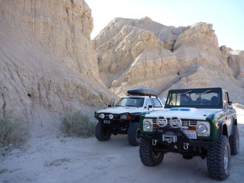 1985 Toyota 4Runner - Two SUVs, a green Ford Bronco and a white Toyota 4Runner, are parked side-by-side in a sandy desert landscape with dramatic rock formations.