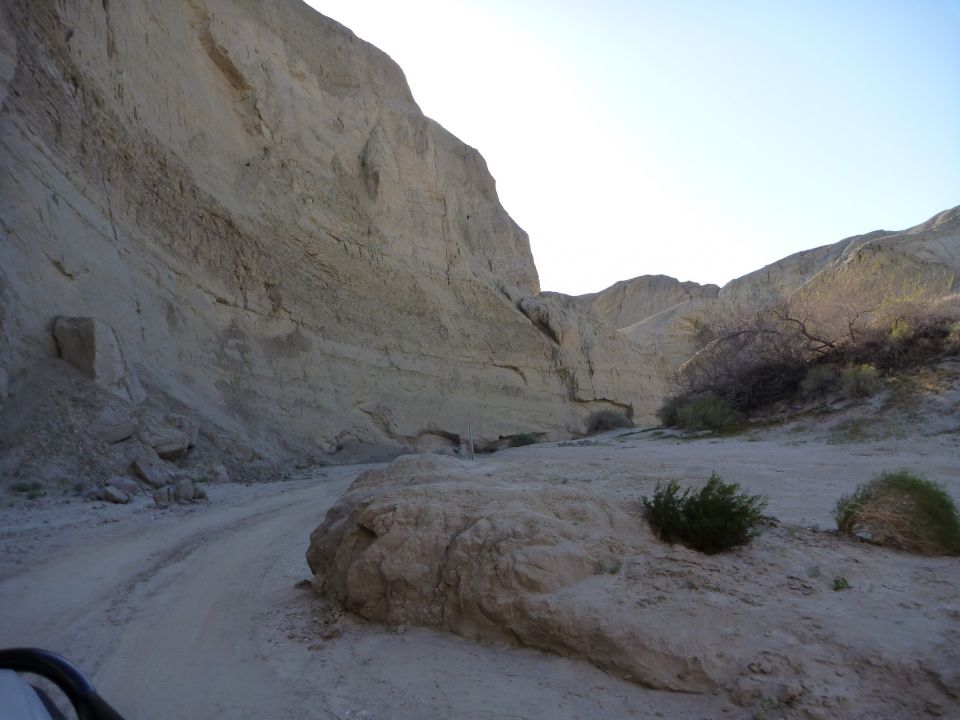 Dirt road winds through a desert canyon with rocky walls and sparse vegetation under a clear sky