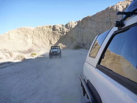 1985 Toyota 4Runner - Two off-road vehicles kick up dust on a dirt path through a desert landscape with steep, eroded canyon walls.