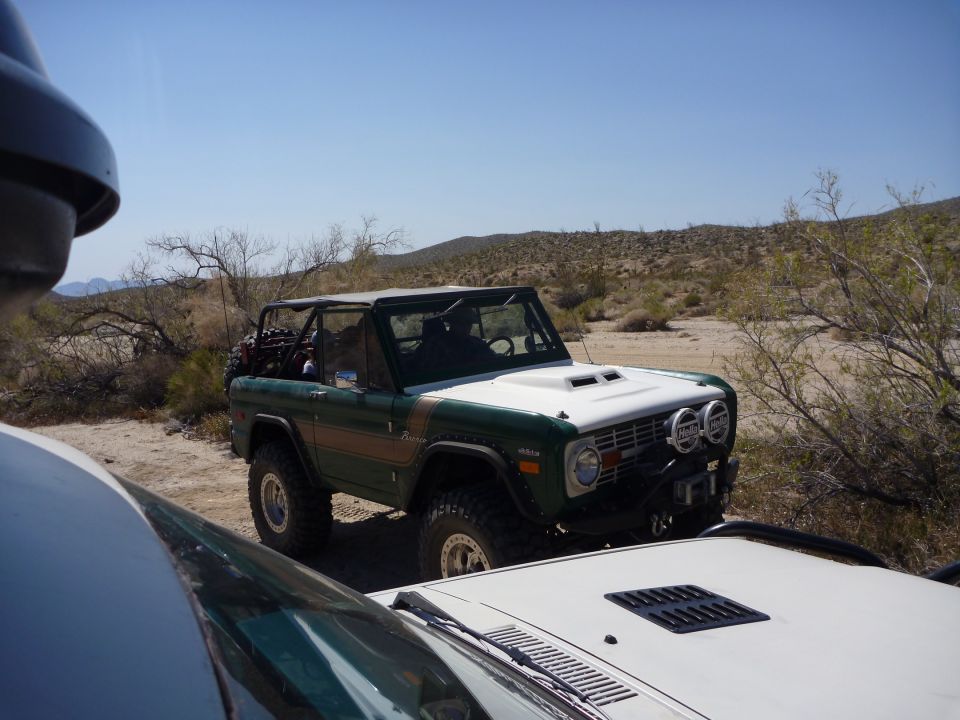 Green and white Ford Bronco with roll cage parked in desert, another vehicle in foreground