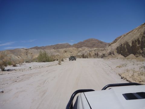 1985 Toyota 4Runner - Two SUVs traverse a dusty desert road, with arid mountains rising in the background under a clear blue sky.