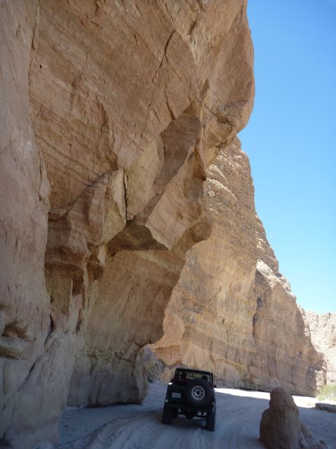 1985 Toyota 4Runner - A dark green SUV navigates a sandy track through a narrow canyon, dwarfed by towering rock walls under a clear blue sky.