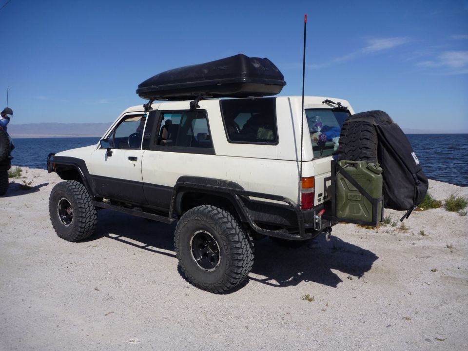 White 1985 Toyota 4Runner with a roof box and spare tire on the back, parked by a body of water