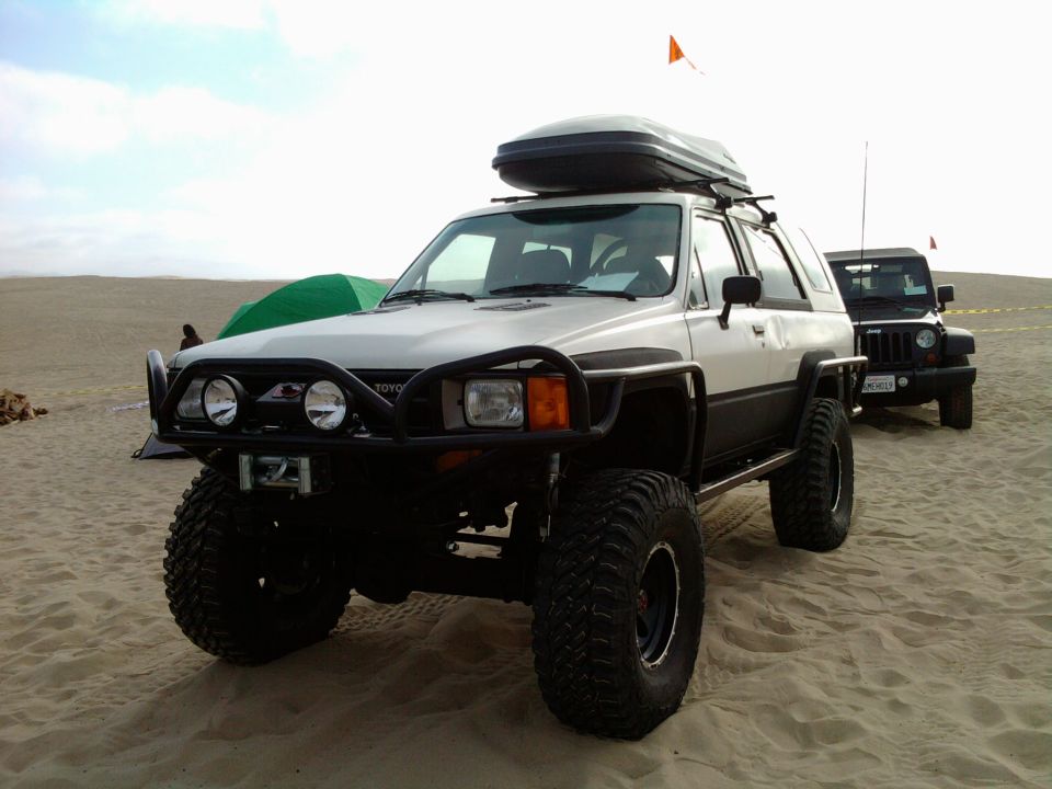 Two-tone 1985 Toyota 4Runner with a roof-top cargo box and off-road tires parked on sand