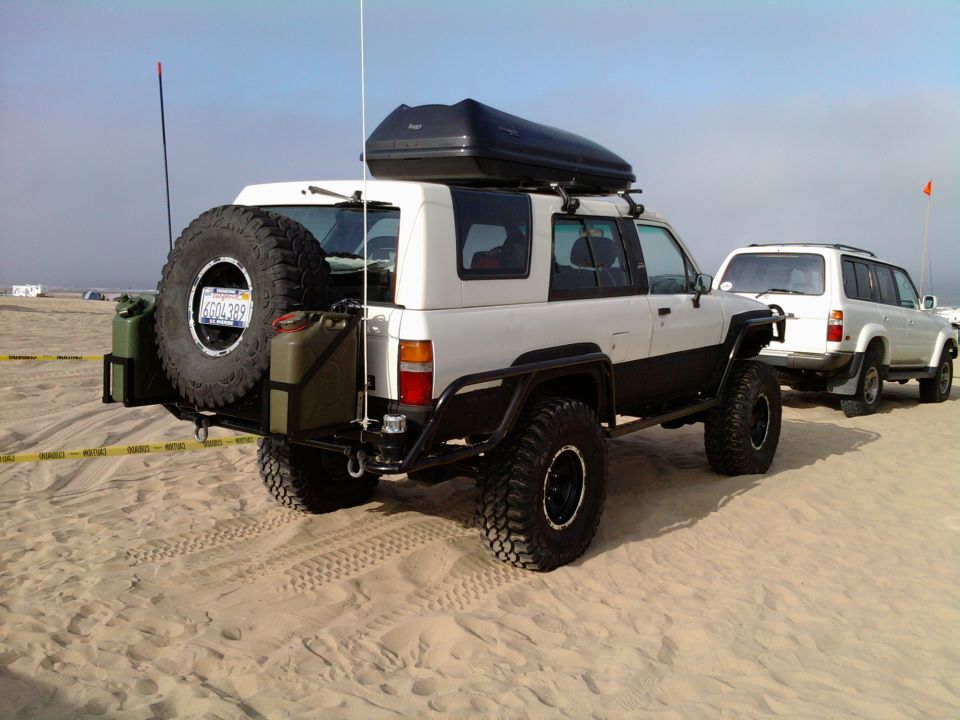 Modified Toyota SUV with spare tire and jerry cans on rear bumper, parked on sand with another SUV behind it