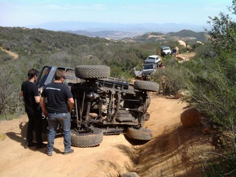 1985 Toyota 4Runner - An overturned 1985 Toyota 4Runner lies on its side on a dusty off-road trail, with other vehicles and onlookers in the distance.