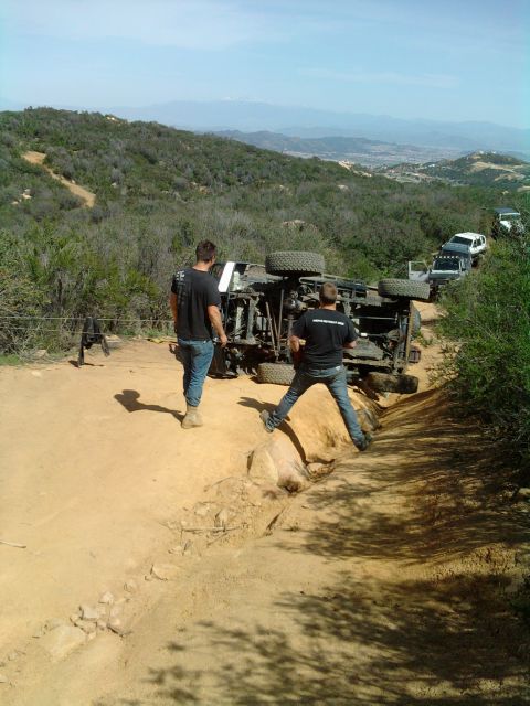 1985 Toyota 4Runner - An off-road vehicle has overturned on a steep dirt trail, with several other vehicles lined up behind it.