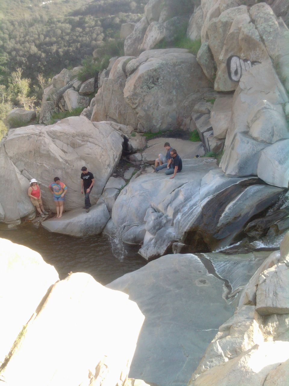 Group of people hanging out by a waterfall in a rocky area. One person is wearing a white hat