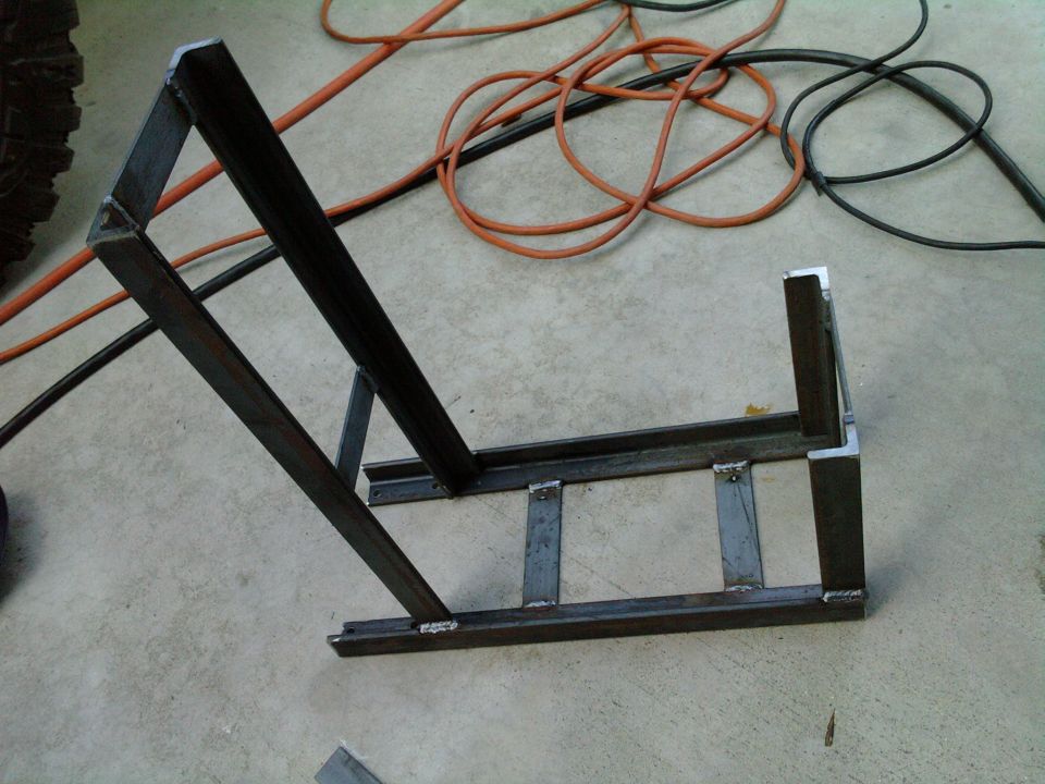 Welded metal frame, possibly for a jerry can carrier, on a concrete floor with orange and black cords