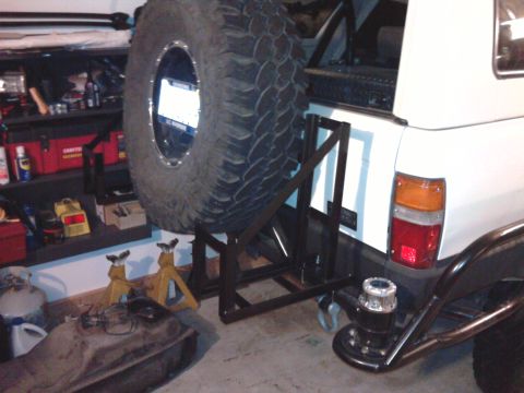 1985 Toyota 4Runner - A custom spare tire carrier is mounted to the rear of a white vehicle, with a fuel tank and jack stands visible in the foreground.