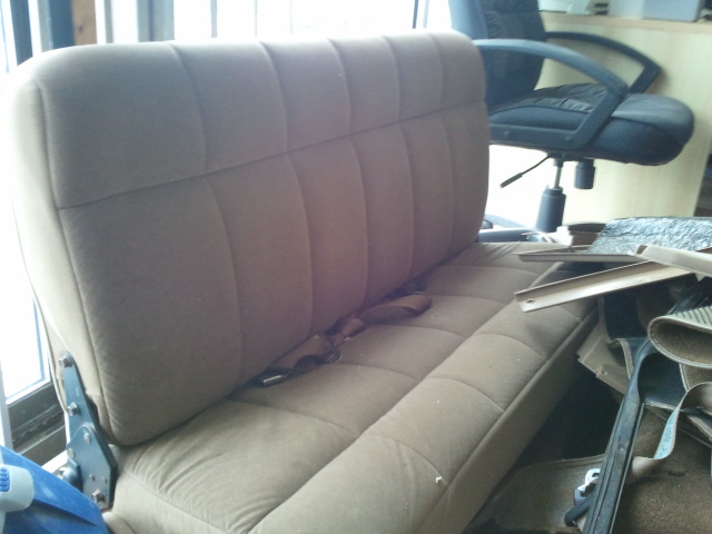 Brown fabric bench seat with seatbelts, in front of an office chair and some clutter