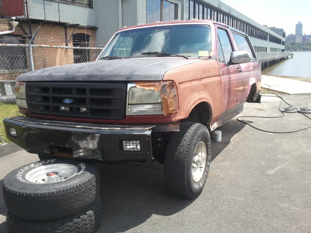Red Ford Bronco with a grey hood and aftermarket bumper, missing front fender, parked outdoors