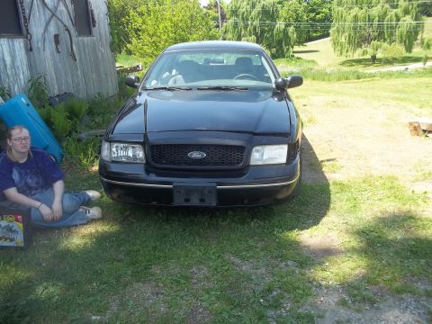 2004 Ford Crown Victoria - A black Ford Crown Victoria is parked in a grassy area next to a building, with a person sitting on the ground to its left.