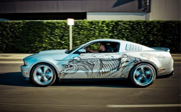 Silver Ford Mustang with custom skeleton graphics driving down a road. It has a GT badge on the fender