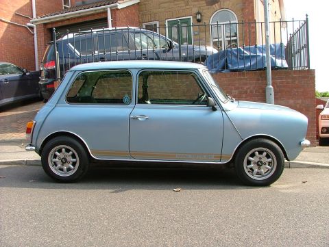 1980 MINI Cooper - A light blue classic Mini Cooper is parked on the street, with other vehicles and a house in the background.