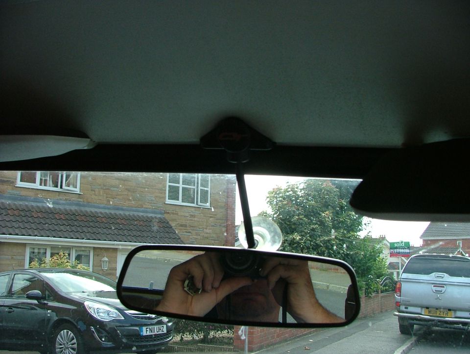 Car interior with rearview mirror reflecting a person holding a camera, showing a dark hatchback and a silver pickup truck outside