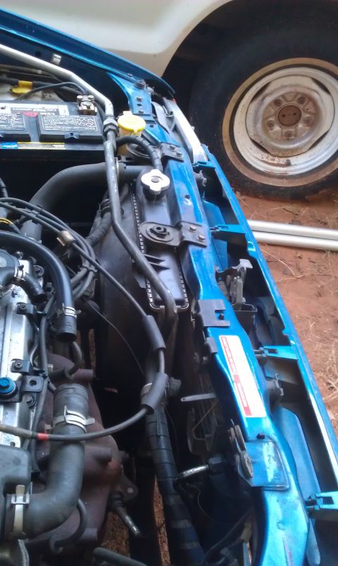 1991 Ford Festiva - Close-up view of a blue car's engine bay, showing the radiator, battery, and various hoses.