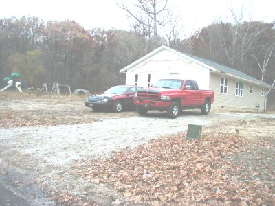 1997 Ford Taurus - A red pickup truck and a dark sedan are parked in a gravel driveway next to a house.