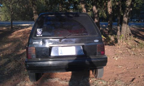 1991 Ford Festiva - Rear view of a black 1991 Ford Festiva hatchback parked on a dirt lot with trees in the background.